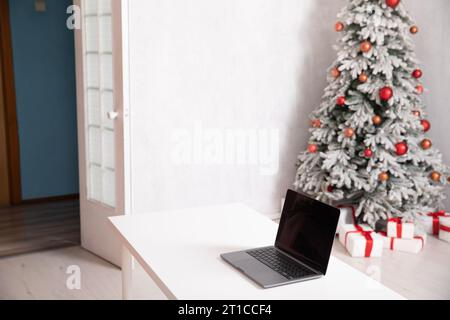 Ordinateur portable sur la table et sapin de Noël décoré avec des cadeaux dans l'intérieur de la chambre blanche Banque D'Images