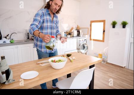 Beau jeune homme barbu caucasien en chemise à carreaux bleus et denim, versant de l'huile d'olive dans un bol blanc avec de la salade fraîche saine. Charmant mâle ch Banque D'Images