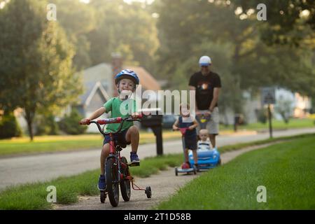 Deux garçons à vélo et scooter ; père pousse bébé dans une voiture, joyeux Banque D'Images