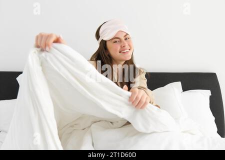 Belle jeune femme avec masque de sommeil sur le front, se réveillant le matin et souriant, se reposant dans sa chambre, se sentant paresseux le week-end Banque D'Images