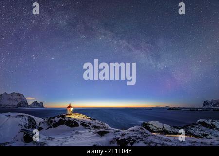 Phare illuminé se dresse sur le promontoire avec des montagnes enneigées et ciel étoilé avec voie lactée, Senja, Troms og Finnmark, Norvège Banque D'Images