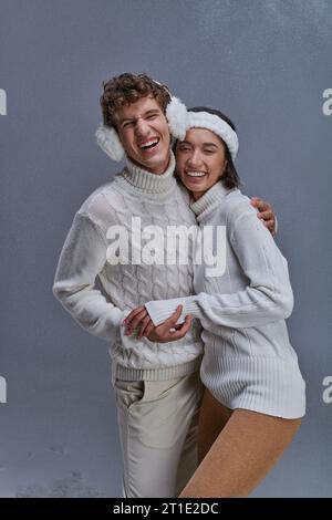 couple interracial en pulls blancs embrassant et riant sur fond gris enneigé, mode hiver Banque D'Images