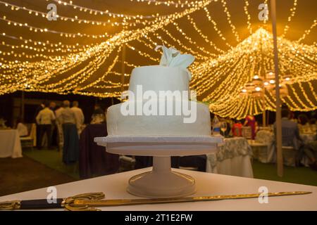 Gâteau de mariage fondant sur la table avec épée dans la salle de banquet. Tente décorée avec guirlandes LED comme arrière-plan Banque D'Images