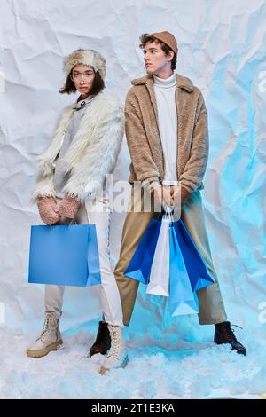 shopping d'hiver, modèles de mode interracial en tenue chaleureuse et confortable avec des sacs à provisions dans le studio enneigé Banque D'Images