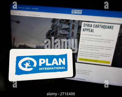 Personne tenant un téléphone cellulaire avec le logo de l'organisation de développement Plan International devant la page Web. Concentrez-vous sur l'affichage du téléphone. Banque D'Images