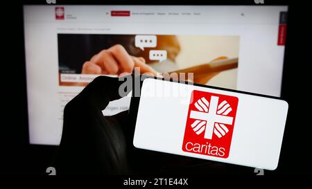 Personne tenant un téléphone portable avec le logo de l'organisation allemande Deutscher Caritasverband e.V. devant la page Web. Concentrez-vous sur l'affichage du téléphone. Banque D'Images