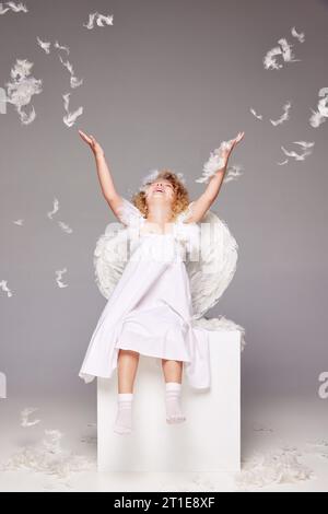 Petite fille heureuse, souriante, positive, enfant en costume d'ange assis isolé sur fond de studio gris avec des plumes volantes Banque D'Images