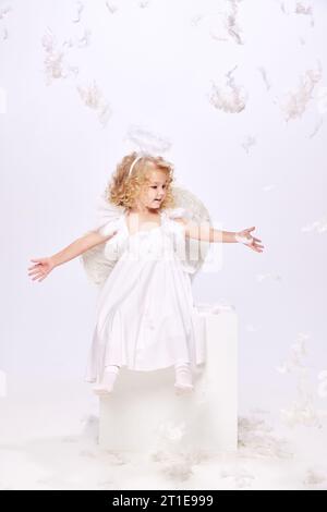 Petite fille heureuse, souriante, positive, enfant en costume d'ange assis isolé sur fond de studio blanc avec des plumes volantes Banque D'Images