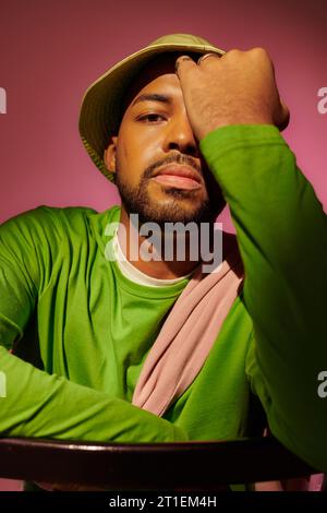beau jeune homme avec la barbe dans le chapeau vert avec la paume sur son visage regardant la caméra, concept de mode Banque D'Images