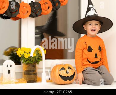 Concept Halloween. Un petit garçon joyeux et beau dans un chapeau de sorcier et un chandail orange est assis sur une table dans la cuisine sur le fond d'un Banque D'Images