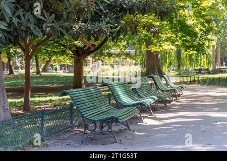 Cinq bancs verts vides dans une rangée dans un parc verdoyant à Paris, France. Dans le fond flou, les gens marchent ou s'assoient dans le parc ensoleillé. Banque D'Images