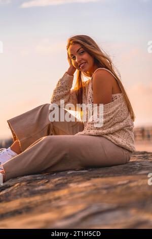 Style de vie d'été, une jeune femme blonde caucasienne assise au bord de la mer dans un haut crop blanc et un pantalon en velours côtelé. Profiter et sourire sur un été Banque D'Images