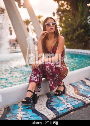 Portrait d'une jeune brune dans une robe florale marron et lunettes de soleil blanches modernes profitant de l'été dans l'heure d'or dans une fontaine de la ville Banque D'Images