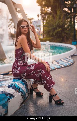 Portrait d'une jeune brune dans une robe florale marron et lunettes de soleil blanches modernes profitant de l'été dans l'heure d'or dans une fontaine de la ville Banque D'Images