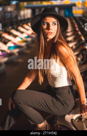 Lifestyle, portrait d'une caucasienne brune avec pantalon long, t-shirt blanc et chapeau noir dans un parc d'attractions dans les voitures tamponneuses Banque D'Images