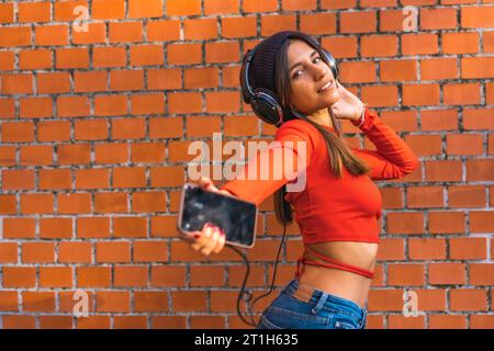 Lifestyle, jeune brune avec un bonnet en laine et un pull rouge profitant de la musique de son téléphone portable avec son casque noir. Fond d'un mur de briques Banque D'Images