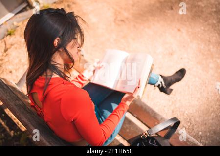 Style de vie. Jeune brunette caucasienne en chemise rouge assise sur un banc dans le parc lisant un livre. Photo détaillée vue du dessus Banque D'Images