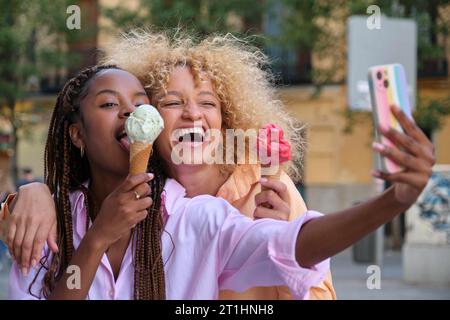 Deux amis heureux multiraciaux avec des glaces prenant un selfie en été dans la rue. Banque D'Images