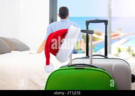 Une valise rouge, un chapeau de Père Noël et un touriste célibataire se relaxant regardant par la fenêtre d'une chambre d'hôtel après être arrivé avec des valises au premier plan Banque D'Images