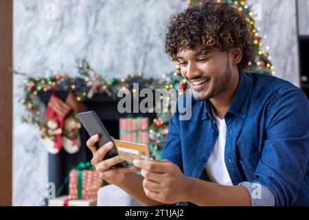 Jeune homme à Noël à la maison dans le salon assis près de l'arbre de Noël décoré avec des décorations de Noël, souriant hispanique tenant une carte de crédit bancaire et téléphone, choisissant des cadeaux en ligne. Banque D'Images