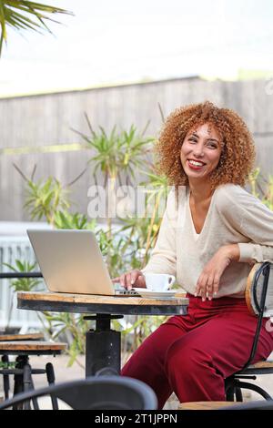 FEMME AUX CHEVEUX BOUCLÉS TRAVAILLANT SUR ORDINATEUR PORTABLE EN PLEIN AIR ET SOURIANT Banque D'Images