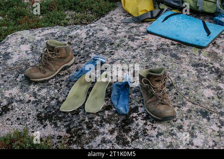 Vieilles bottes de trekking brunes, semelles vertes et chaussettes bleues à sécher après une journée de randonnée, disposées sur pierre. Pieds mouillés en voyage. Camping de voyage Banque D'Images