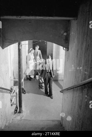 Guy's Hospital- La vie dans un hôpital de Londres, Angleterre, 1941 Une infirmière accompagne deux porteurs comme ils le transport d'un patient dans la salle d'opération dans le sous-sol de Guy's Hospital à travers les souterrains de l'escalier qui mène jusqu'à la tutelle. Le patient semble être allongé sur un chariot de l'hôpital. Banque D'Images