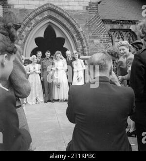 Curé- les travaux de la vicaire de St Mark's Church, Victoria Docks, Silvertown, Londres, Angleterre, RU, 1944, un couple de jeunes mariés pose devant l'appareil photo lorsqu'ils quittent l'église St Mark's, Silvertown après leur mariage. Photographe de leur mariage peut être vu dans cette photographie, comme plusieurs de leurs invités à regarder la procédure. Banque D'Images