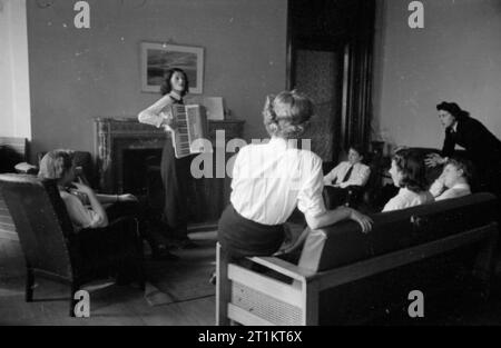 La jeune fille inconnue derrière la bataille de la mer- le travail de Women's Royal Naval Service, 1942 Wren inconnu, son dos à la caméra, et ses collègues profitez d'une soirée musicale dans le salon. L'une des "Roitelets" est à jouer de l'accordéon par la cheminée, tandis que d'autres vous détendre sur les canapés et chanter. Banque D'Images