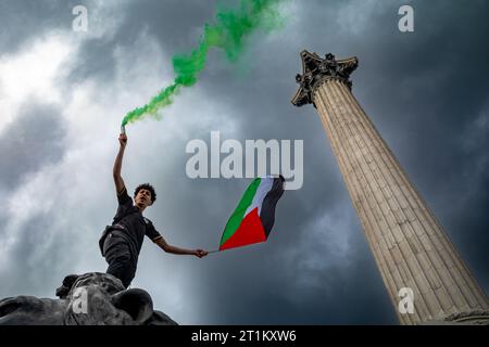 Londres, Royaume-Uni. 14 octobre 2023 : un manifestant pro-palestinien se tient debout sur un lion géant près de la colonne de Nelson tenant une fusée éclairante verte et un drapeau palestinien dans une manifestation en soutien à la Palestine et contre les attaques israéliennes sur Gaza. Banque D'Images