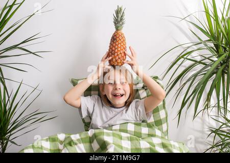 Un garçon de 9 à 10 ans, ses cheveux tombant en cascade, s'allonge dans son lit, tenant doucement un ananas, incarnant l'essence de la détente et des loisirs Banque D'Images