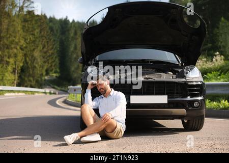 Homme stressé assis sur la route asphaltée près de la voiture cassée à l'extérieur Banque D'Images