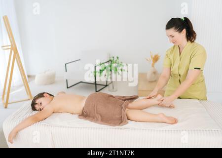 Vue latérale d'un garçon de cinq ans allongé sur une table de massage ayant un massage de drainage lymphatique des jambes et du bas des jambes. Portrait de massothérapeute féminine Banque D'Images