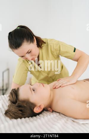 Portrait vertical en gros plan d'un ostéopathe féminin ajustant la poitrine d'un garçon de cinq ans allongé sur une table de massage. Petit enfant en physiothérapie Banque D'Images