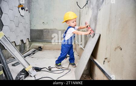 Enfant dans le casque de construction de sécurité vérifiant la surface du mur avec le niveau à bulle dans l'appartement. Enfant dans la combinaison de travail en utilisant l'instrument de niveau tout en travaillant sur la rénovation de la maison. Banque D'Images