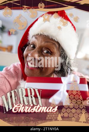 Joyeux texte de noël avec des décorations en or sur femme biracial senior souriante dans le chapeau de père noël Banque D'Images