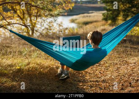 Jeune femme dans hammok à l'aide d'un ordinateur portable travaillant à l'extérieur face au lac sur un coucher de soleil. Concept d'opportunité d'emploi à distance Banque D'Images