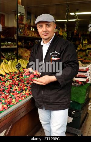 Portrait d'un vendeur de fruits tenant un punnet de papier de baies sauvages. Rangées de fruits frais exposées dans un petit magasin local de la rue Desaix, Paris, France. Banque D'Images