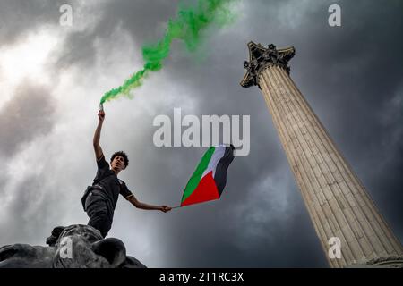 Londres, Royaume-Uni. 14 octobre 2023 : un manifestant pro-palestinien se tient debout sur un lion géant près de la colonne de Nelson tenant une fusée verte et un drapeau palestinien dans une manifestation Banque D'Images