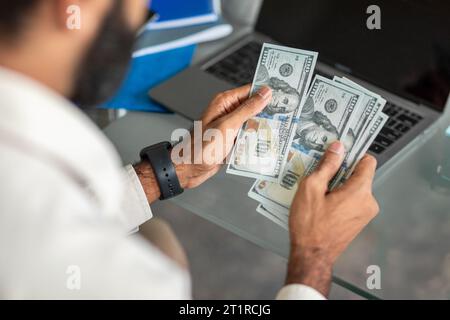 Mains de l'homme d'affaires indien comptant les billets de banque en dollars, assis à la table devant l'ordinateur portable dans le bureau, vue arrière Banque D'Images