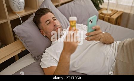 Jeune homme hispanique troublé couché malade en pyjama sur son lit, regardant anxieusement son smartphone tout en tenant une bouteille de pilules dans sa chambre. Banque D'Images