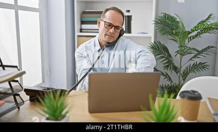 Homme d'affaires d'âge moyen détendu s'engageant dans une conversation téléphonique sérieuse tout en travaillant sur son ordinateur portable au bureau Banque D'Images