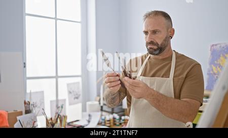 Jeune homme sérieux, un artiste talentueux, se concentrant sur le choix du pinceau parfait dans l'intérieur confortable de son atelier d'art. Banque D'Images