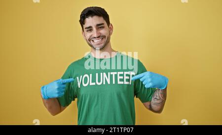 Jeune homme hispanique portant des gants pointant vers l'uniforme bénévole souriant sur fond jaune isolé Banque D'Images