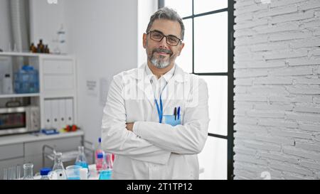 Souriant, confiant, jeune scientifique hispanique aux cheveux gris, debout avec un geste de bras croisés dans le labo Banque D'Images