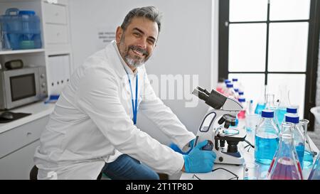 Jeune scientifique hispanique confiant avec les cheveux gris, souriant alors qu'il travaille avec un microscope dans le laboratoire, faisant des découvertes médicales Banque D'Images