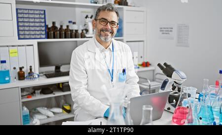 Jeune hispanique séduisant, aux cheveux gris, un scientifique confiant, sourit tout en effectuant une analyse biologique avec son ordinateur portable dans un laboratoire animé. Banque D'Images