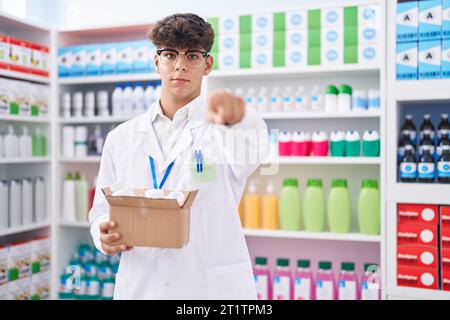 Adolescent hispanique travaillant à la pharmacie pharmacie tenant la boîte avec des pilules pointant avec le doigt à la caméra et à vous, geste confiant regardant seriou Banque D'Images