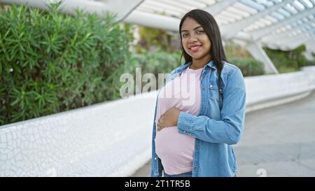 Joyeuse jeune femme latine, attendant joyeusement la maternité, touchant avec confiance son ventre enceinte, souriant dans la lumière du soleil, debout tranquillement au milieu de la ville Banque D'Images