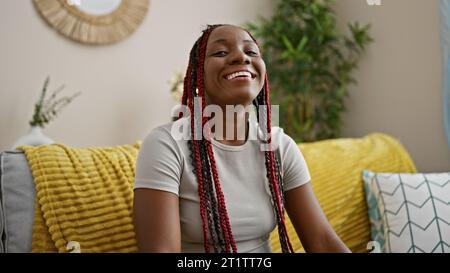 Une femme afro-américaine confiante rayonne de joie, reposant confortablement sur son canapé confortable à la maison, son sourire débordant de positivité et de rires. Banque D'Images